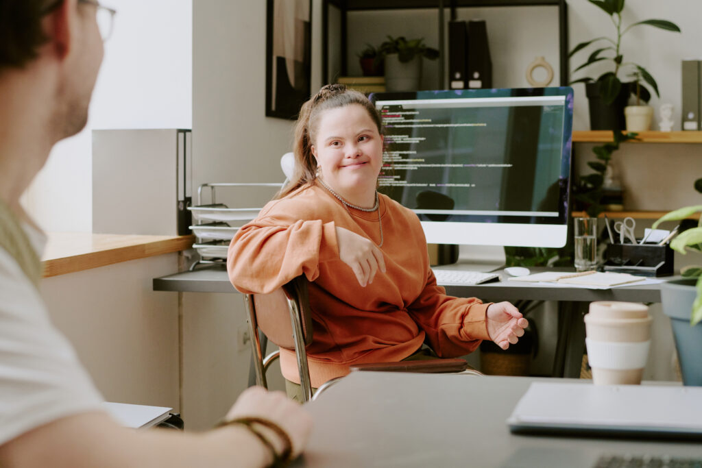 Wide shot of female programmer with down syndrome sitting on chair and looking at her colleague who is out of frame, showing how the Back To Work scheme is inclusive.